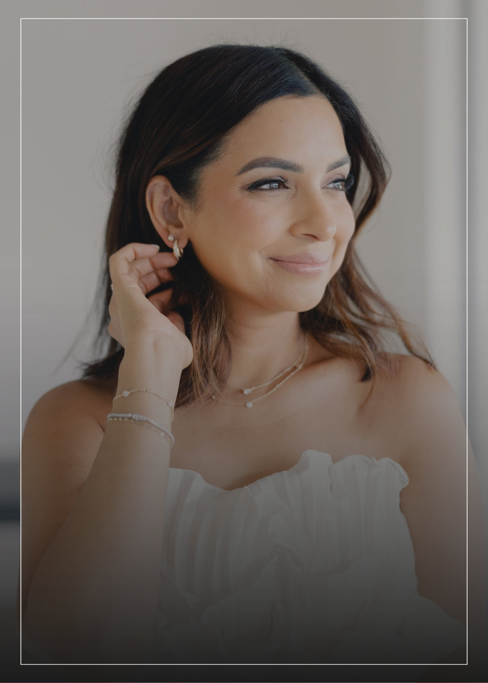 Two smiling women standing close together, one wearing a white, off-shoulder ruffled dress and the other in a beige sleeveless vest over a matching top and white pants, both adorned with delicate gold jewelry, including necklaces and bracelets, in a bright, modern setting.