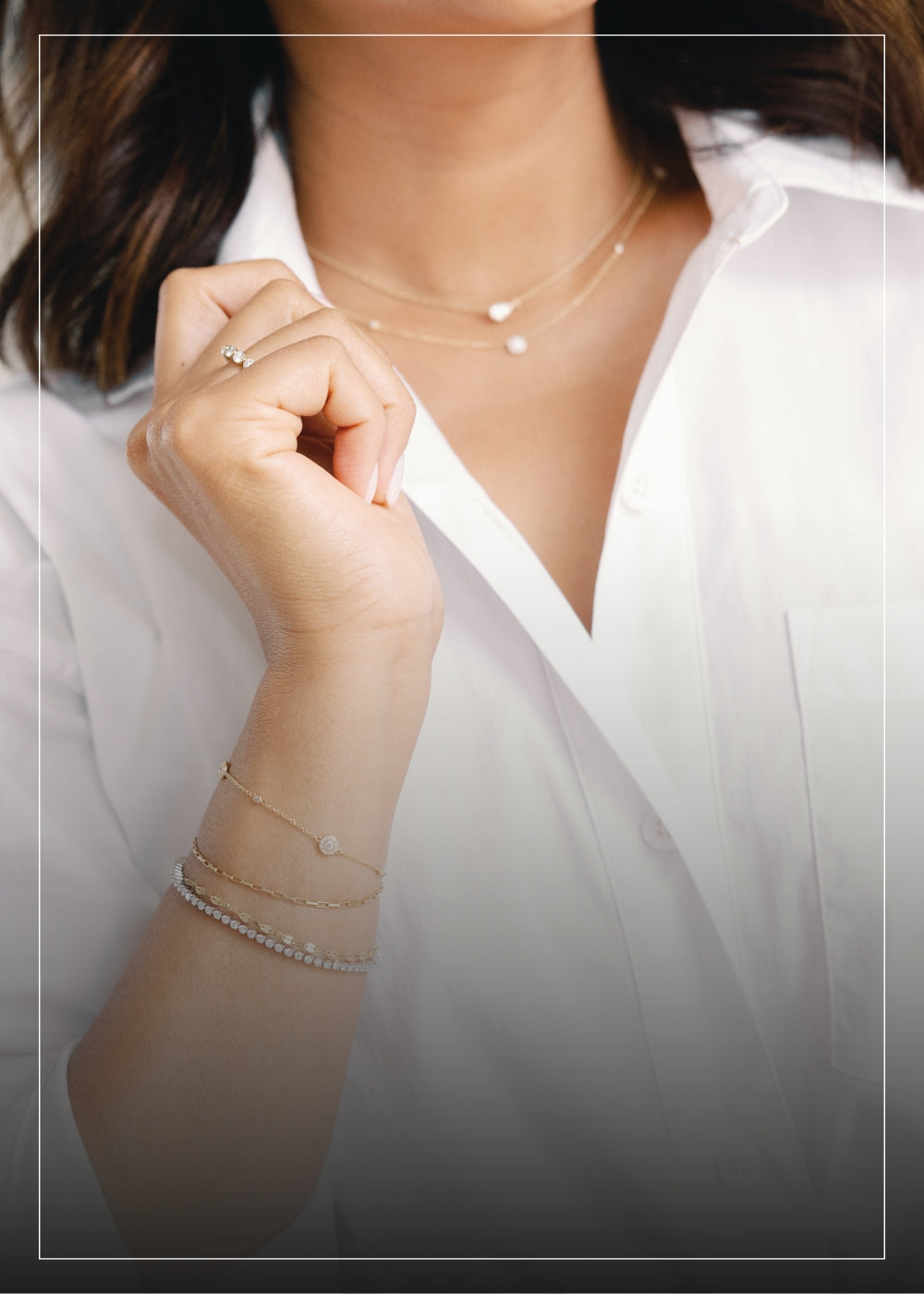 A close-up of a woman's hand and neck, showcasing layered gold necklaces, a gold ring, and multiple gold bracelets, including a diamond-studded bracelet, against the backdrop of a white button-down shirt.