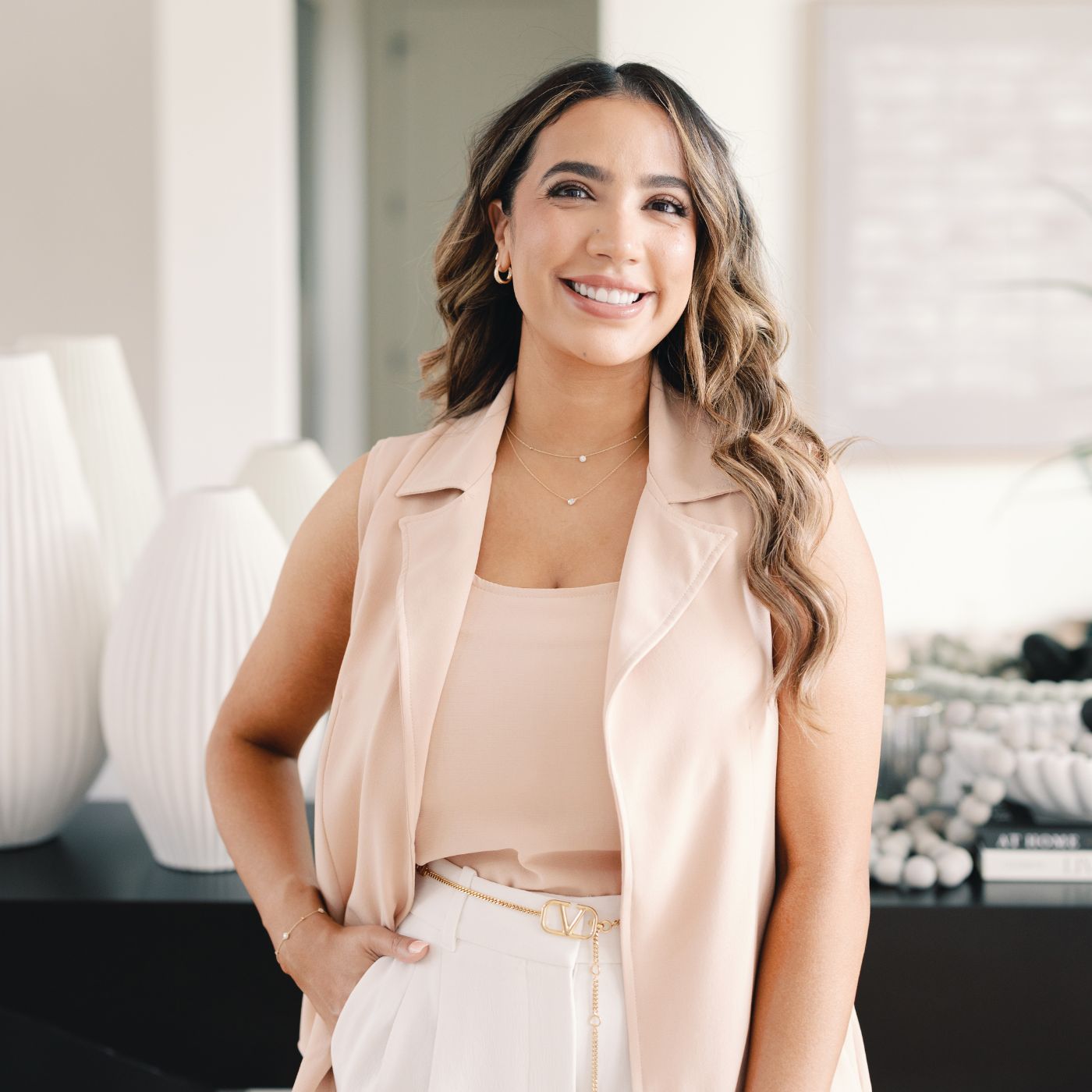 Seema of Seema Jewelry & Co wearing a sleeveless beige vest over a matching top and white pants. She is accessorized with delicate gold jewelry, including a necklace and earrings, standing in a modern, well-lit room with white decorative elements in the background.